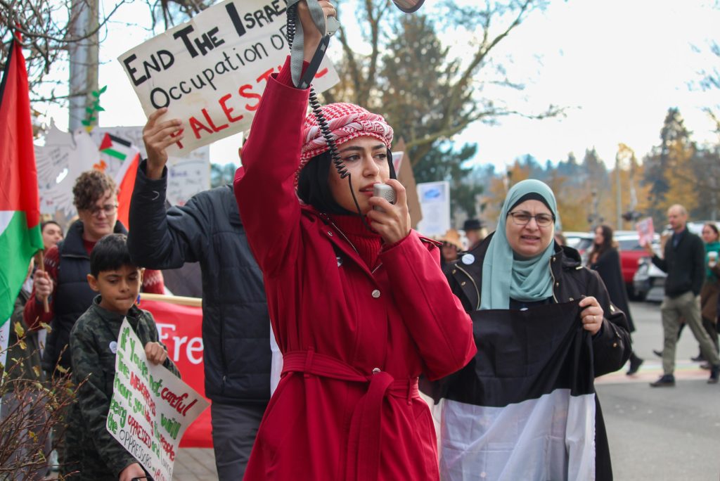 Sara Kishawi holds an attachment, connected to a megaphone that she waves in the air with another hand, as she speaks into it and walks. Behind her are other community members, marching with signs. One of the signs says "End the Israel occupation of Palestine." Sara wears a Keffiyeh scarf on her head.