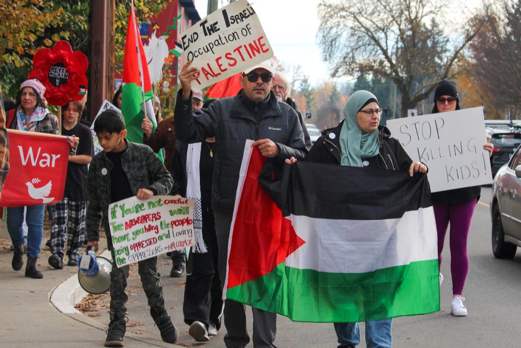 Community members march on the sidewalk and partially on the road, holding the Palestinian flag and signs that read "end the Israel occupation of Palestine," "Stop killing kids!" and "If you're not careful, the newspapers will have you hating the people being oppressed and loving the oppressors - Malcolm X."