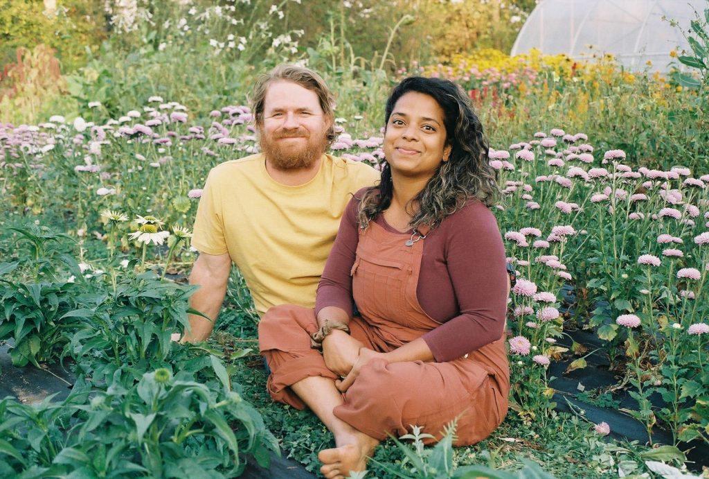 The owners of Wild Bee Florals, Aaron (left) and Thanushi (right) sit in a field surrounded by flowers.