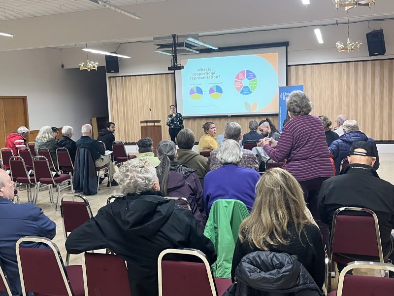 A group of people sit in chairs, photo is taken from behind as they listen to politician Lisa Marie Barron, at the front of the room, during a downhill discussion