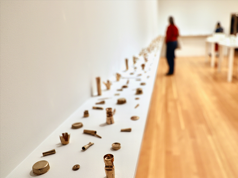 A variety of plastic objects like bottle cap lids and pen lids are cast into bronze and are strewn down a white perimeter ledge in a gallery