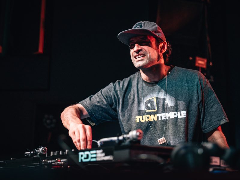 Dj Peter Poole stands in front of two turntables in a ball cap and a T-shirt that reads Turntemple