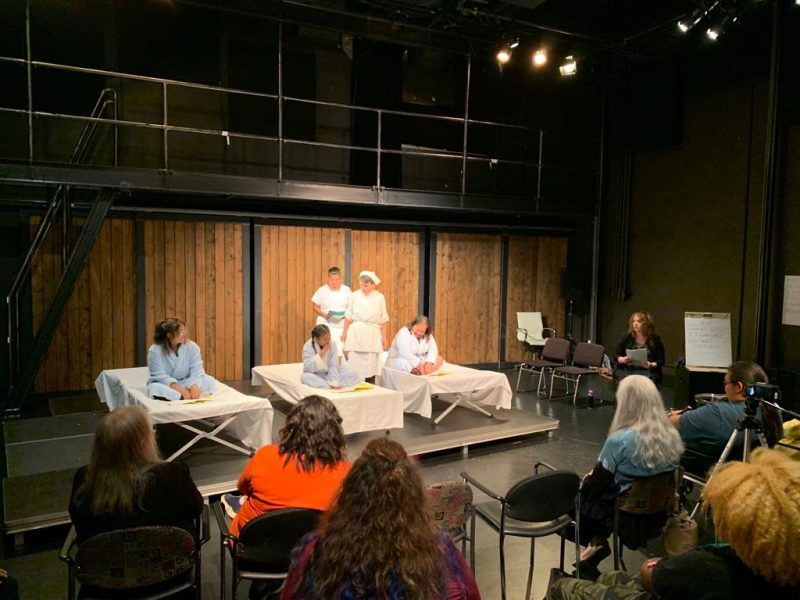 Three young women in robes are onstage and sitting on what appears to be hospital beds as an audience looks on