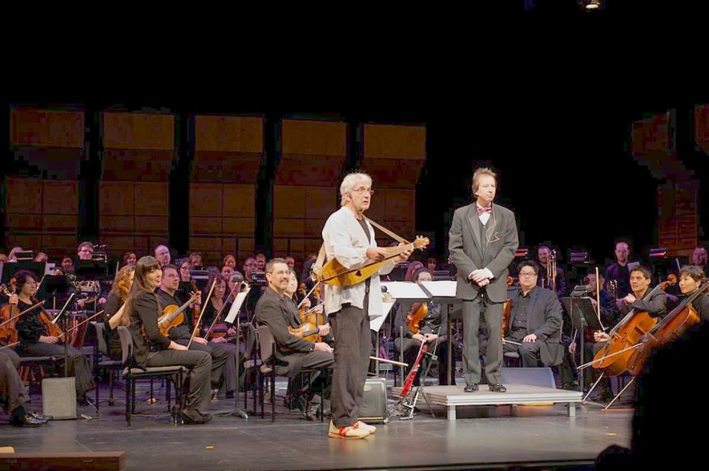 Rick Scott plays the dulcimer onstage beside conductor Pierre Simard. Both men stare out at the audience from the stage, looking a bit confused