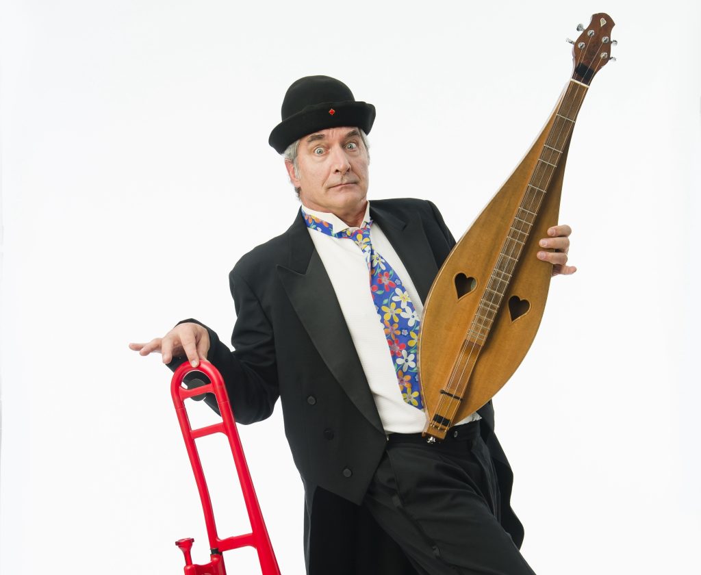 Rick Scott wears a black bowler-style hat and holds a dulcimer in one hand and a red trombone in the other, with a flowered tie