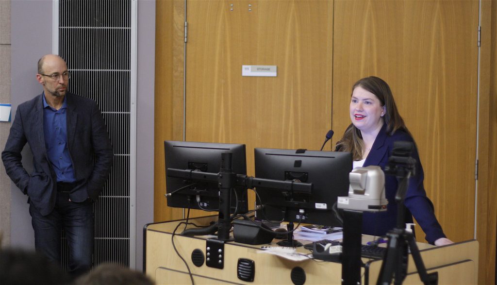 Jen Gerson, a young woman with light brown hair and red lipstick speaks from behind a computer monitor and microphone. David Livingstone, a bald man in suit and glasses, stands to the left and watches