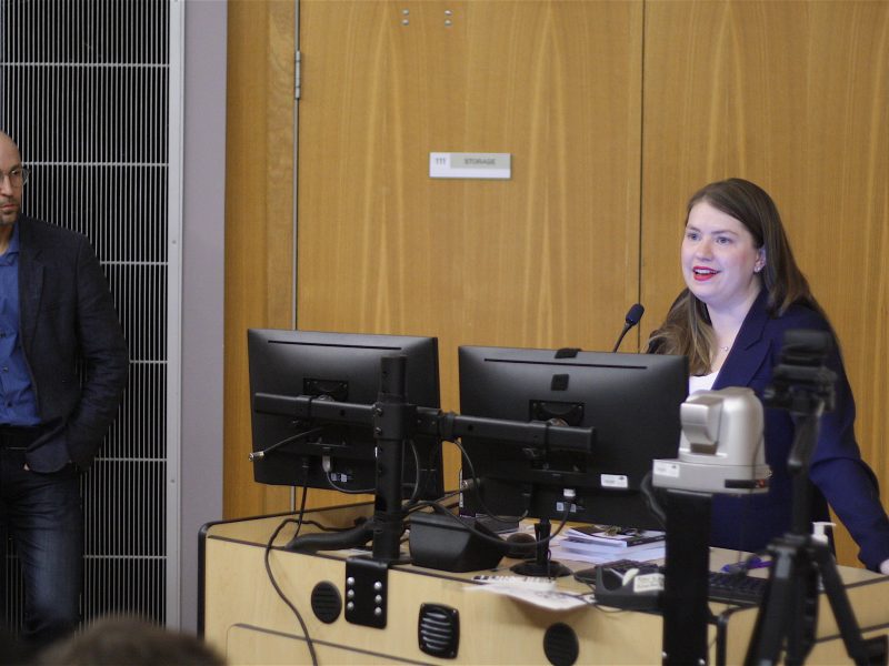 Jen Gerson, a young woman with light brown hair and red lipstick speaks from behind a computer monitor and microphone. David Livingstone, a bald man in suit and glasses, stands to the left and watches