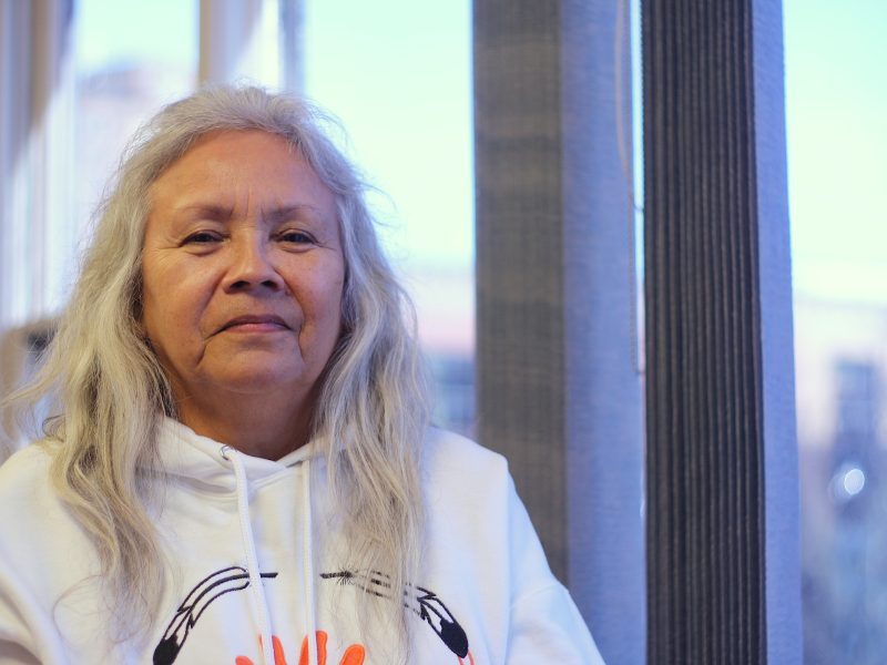 An Indigenous woman with long grey hair and a white hoodie with embroidered feathers looks into the camera
