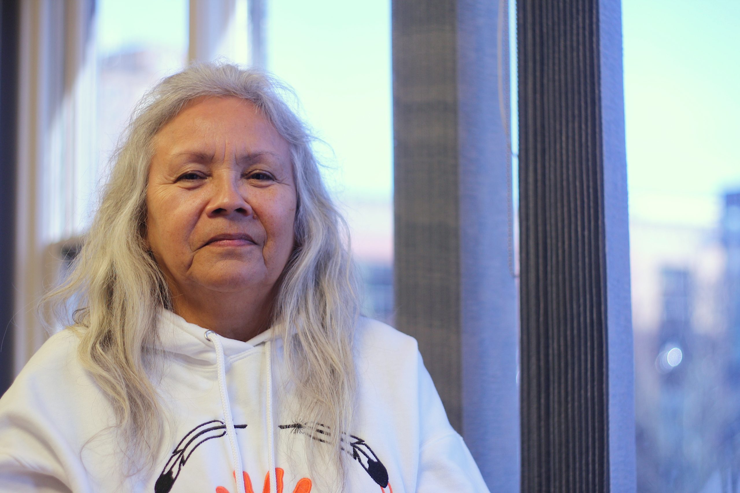 An Indigenous woman with long grey hair and a white hoodie with embroidered feathers looks into the camera