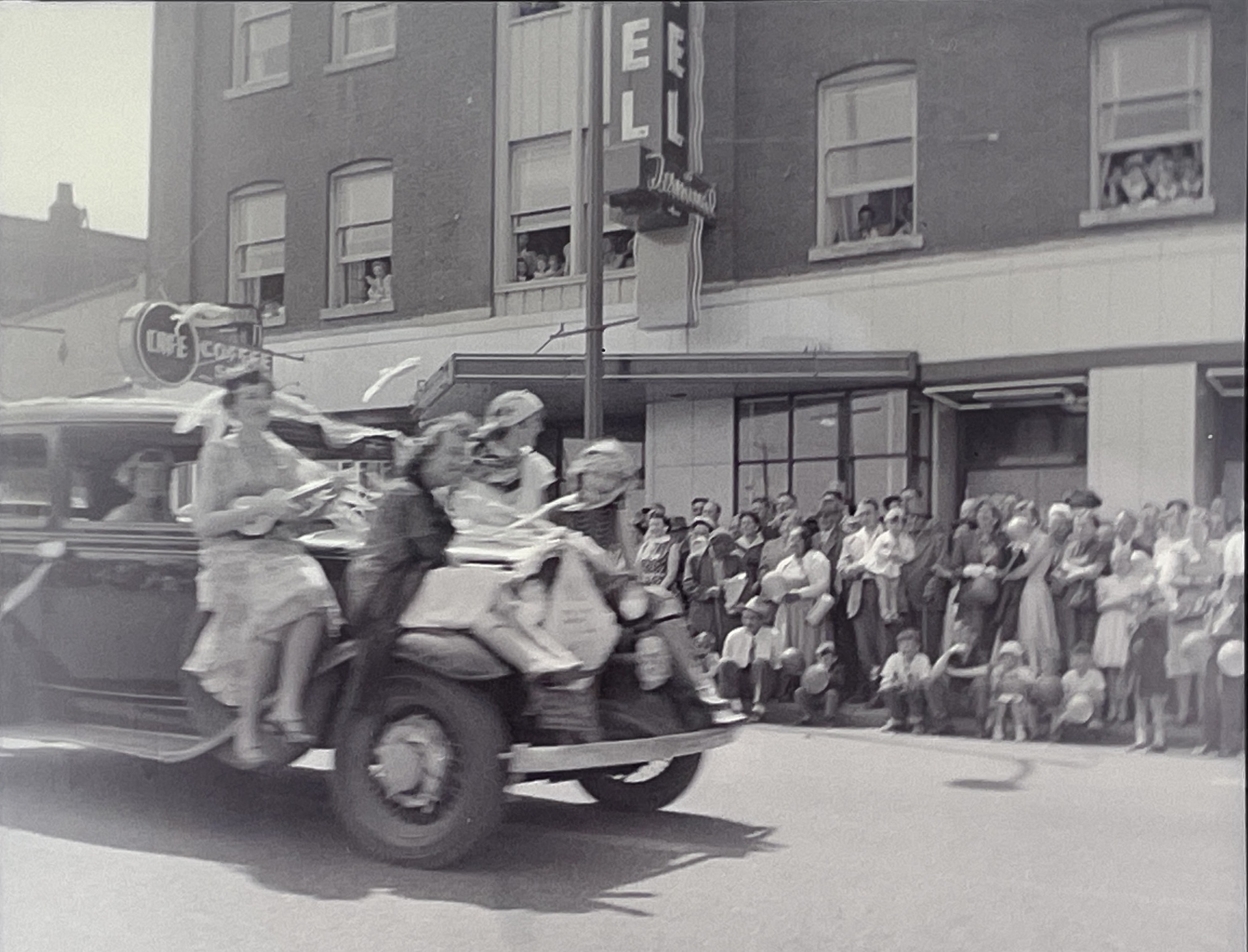 A black and white vintage image from the 1950s shows a blurry image of women on the hood of a car during a parade in downtown Nanaimo