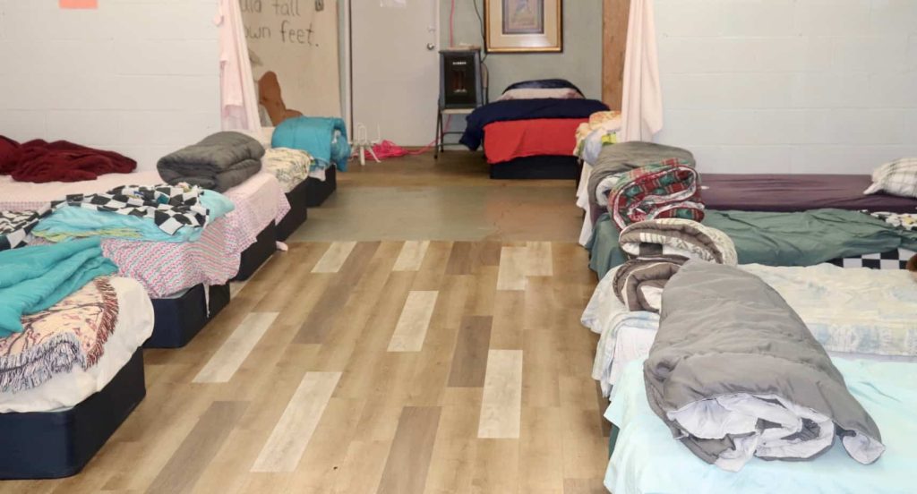 Twin size beds, 12 in total, line both sides of a room. Each bed has different bedding and blankets on it.