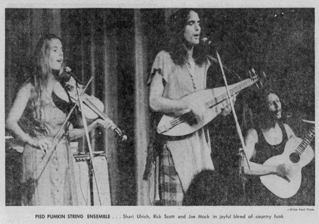 An old black and white newspaper photo shows three musicians from the Pied Pumkin band, with Rick Scott playing dulcimer with long hair at the centre