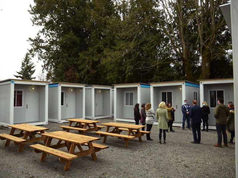 Modular single-suite housing units are arranged in a square formation, all facing inward. In the centre are picnic benches and a group of people standing near one of the units.