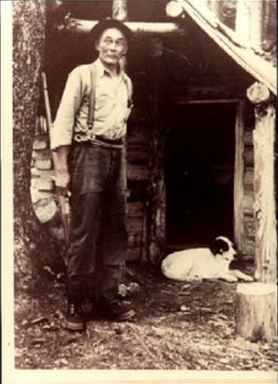 Old Black man stands outside a woodshed. Dog sits next to him.