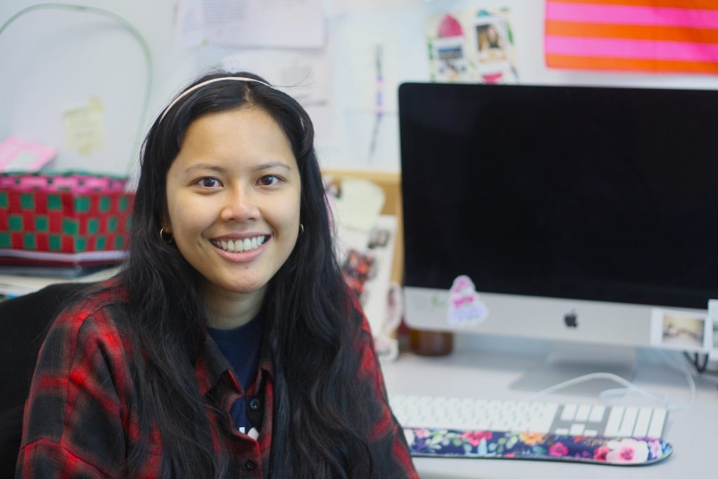A young woman, Sarah Mei Lyana, with long black hair and a black and red checkered shirt, sits at a computer