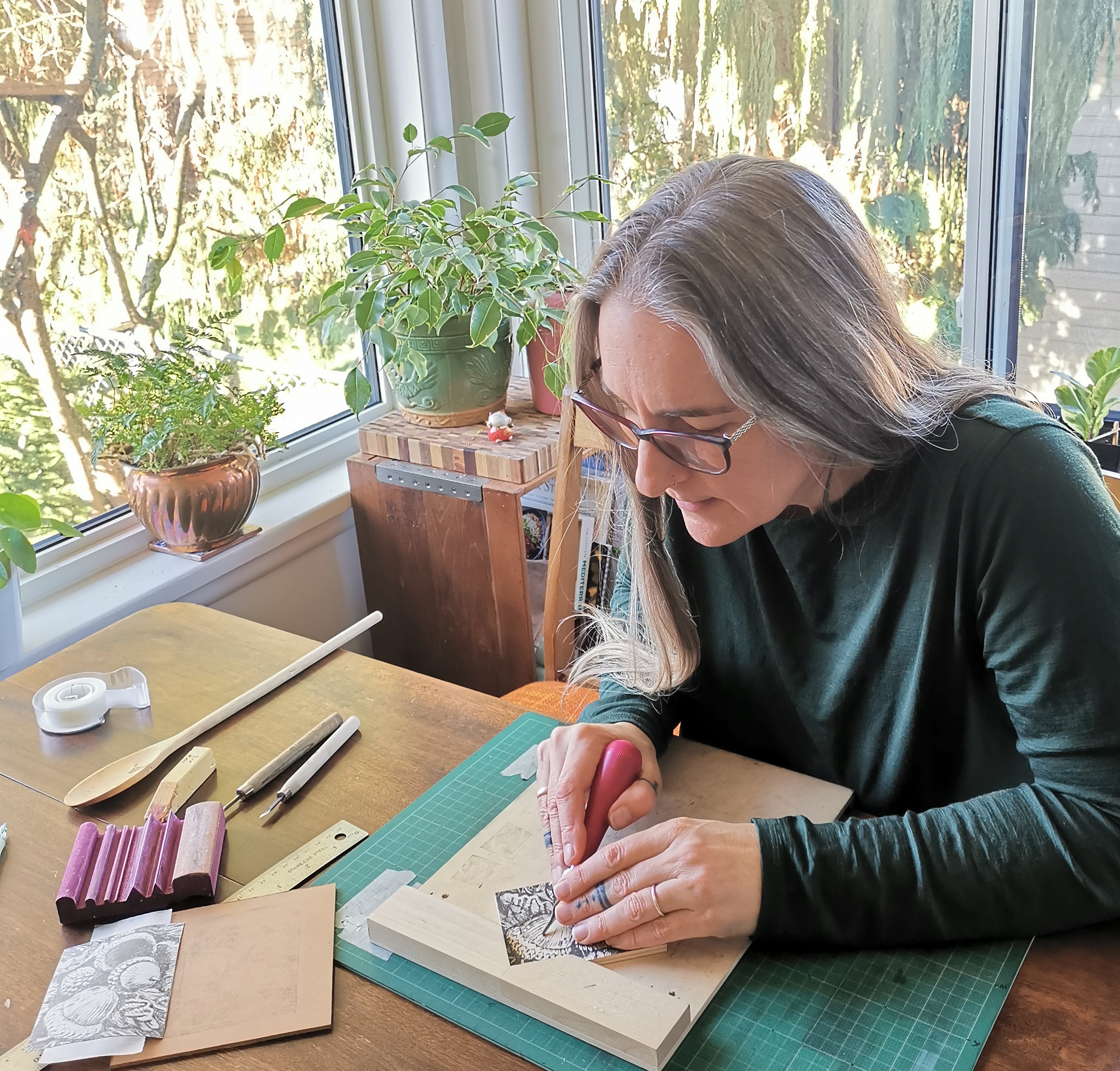 Kerri Orser sits at a sunny table with a variety of linocut tools around her, carving a piece with acorns on it