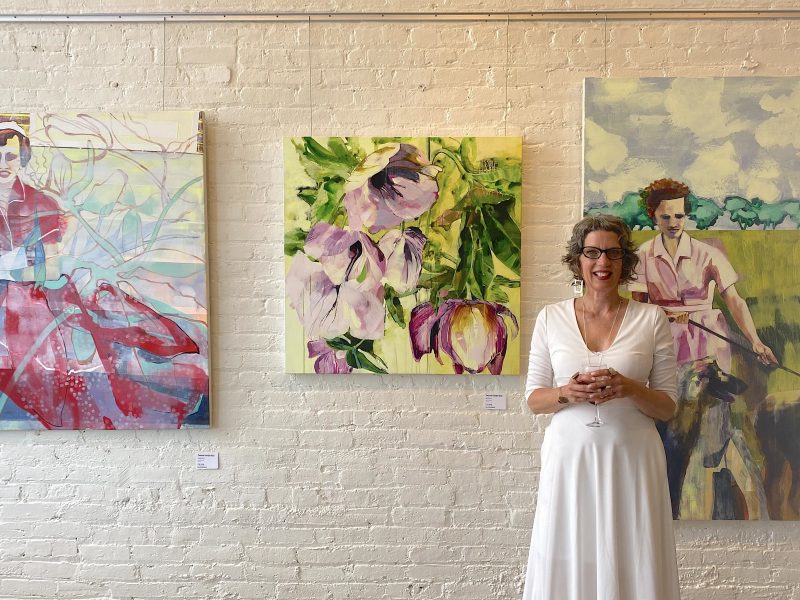Artist Yvonne Vander Kooi stands in front of a collection of large paintings in a white dress