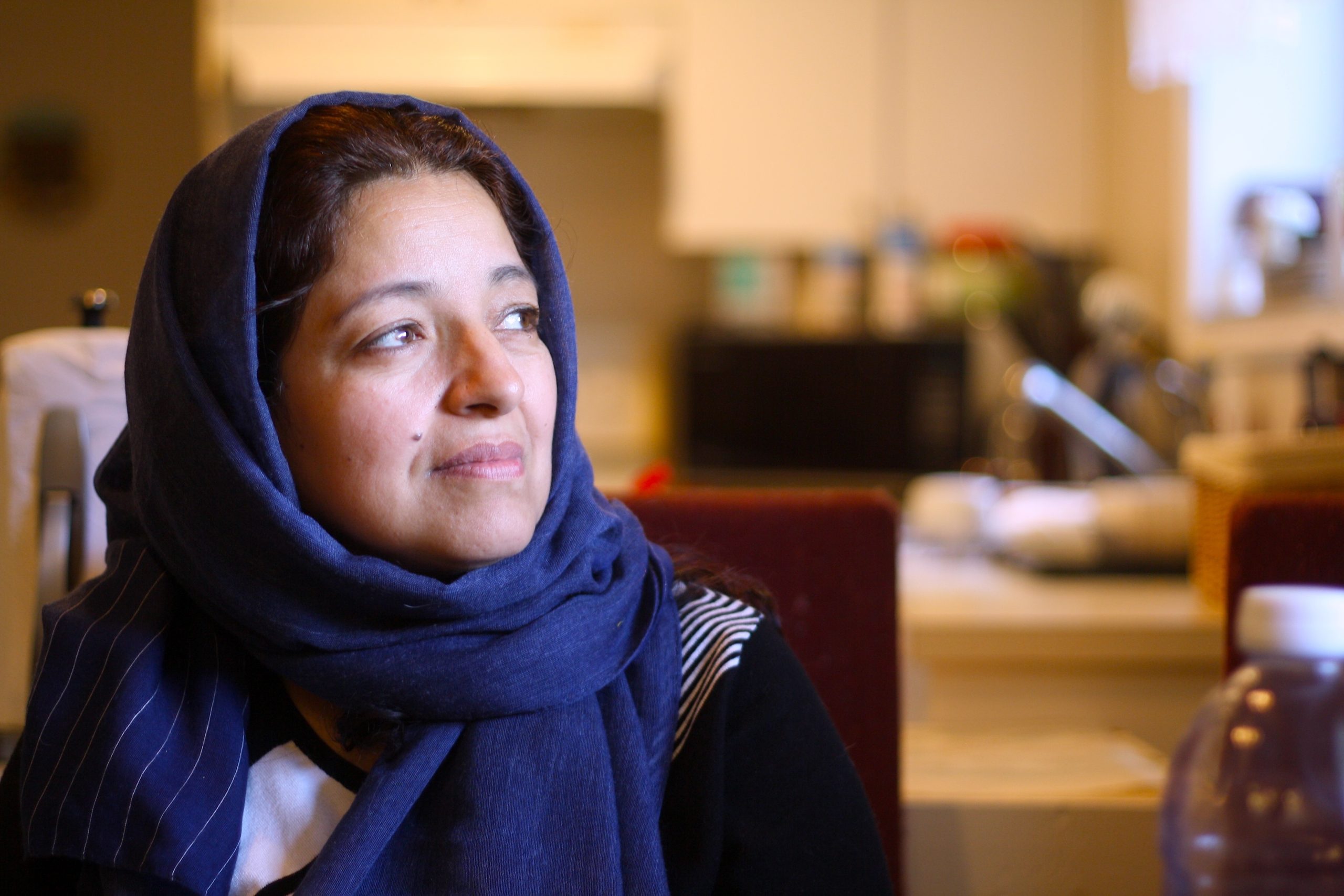 A woman in a blue headscarf sits in her kitchen and looks out the window