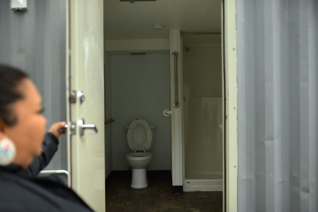 A person opens a door to reveal the inside of the bathroom building at The Village. There is a toilet and standing shower inside.
