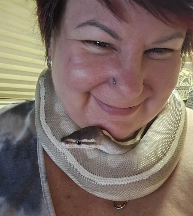 A woman with short brown hair wears a light brown snake around her neck and is smiling