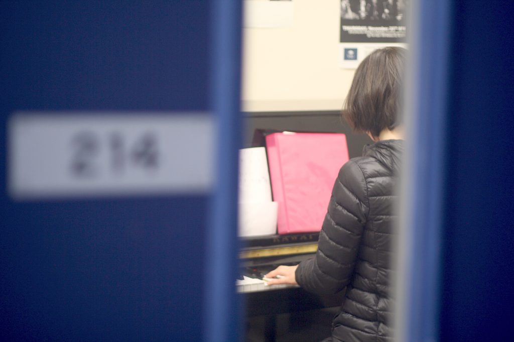 A woman plays piano in a music room, as seen through a window in a door