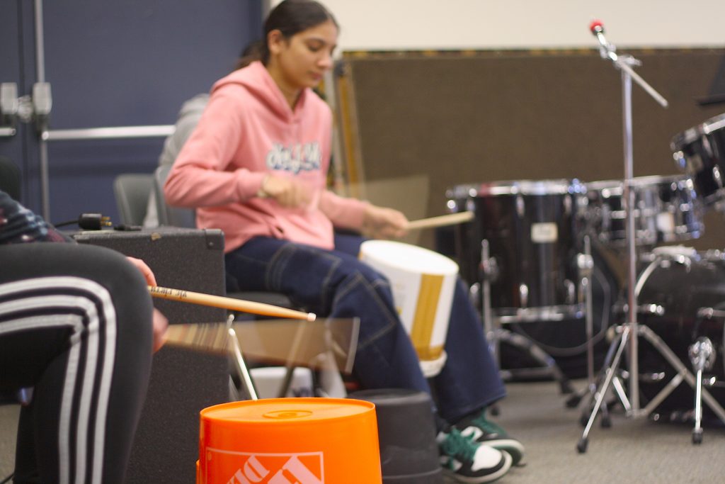 Students practice with drumsticks on orange plastic tubs during a percussion music class