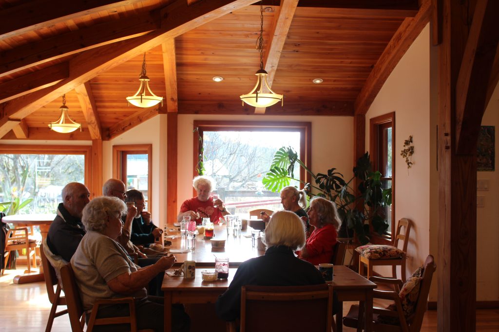 A group of seniors sit in a timber-frame dining room around a large wood table. Sun shines from large windows behind them. They are all enjoying tea.