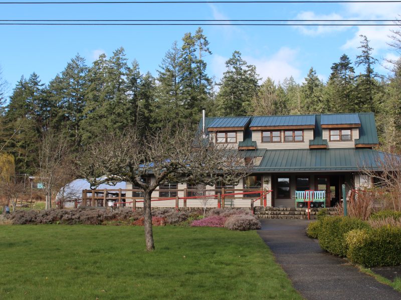 An image of the St. Ann's Garden Club building. A large timber-frame house-like structure with a green roof. In front is a grassy field with a heritage apple tree.