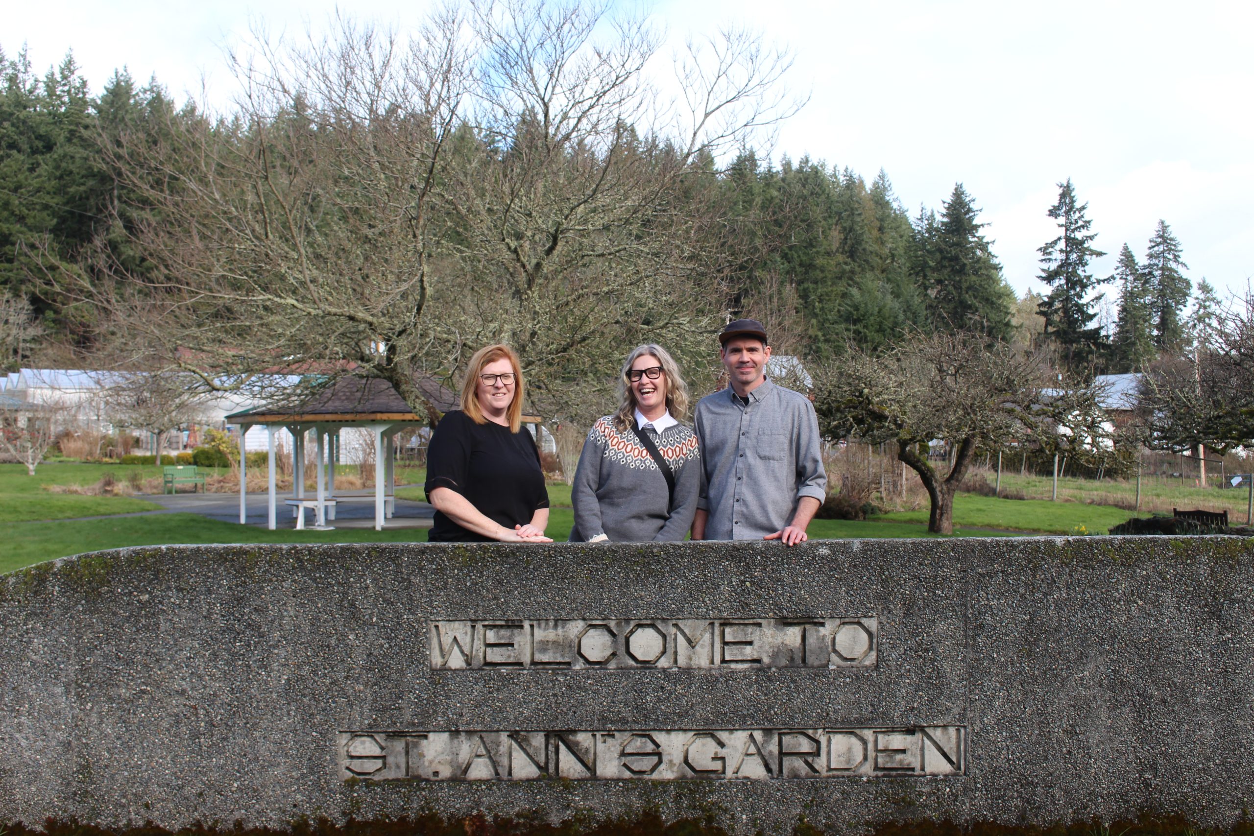 Leah Boisvert, Stephanie Kok and Conrad Murphy stand behind a cement half-wall that has St. Ann's Garden Club engraved in it. Behind them is a field with walking paths and apple trees.