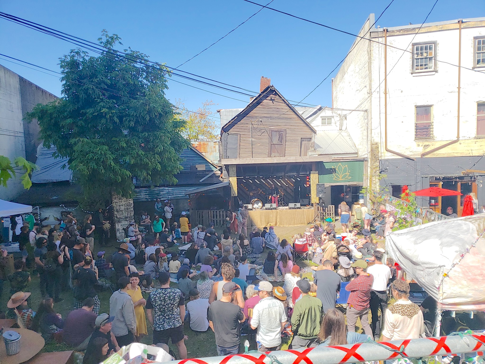 A crowd of audience members gather in a backyard around a makeshift stage constructed of wood