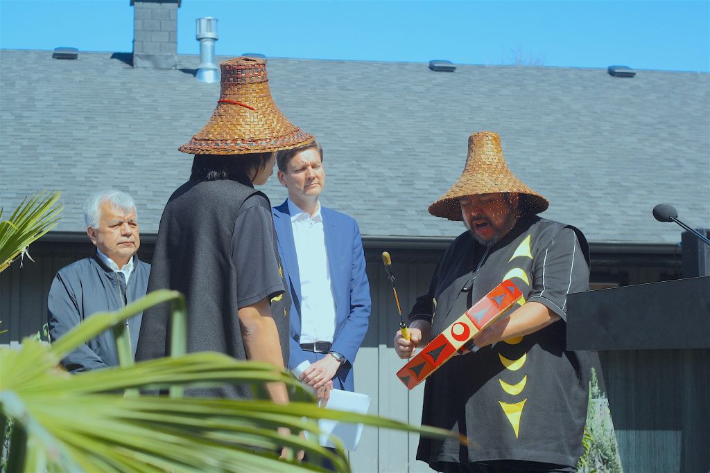 A man in a woven cedar hat songs and drums as Premier Eby and others watch