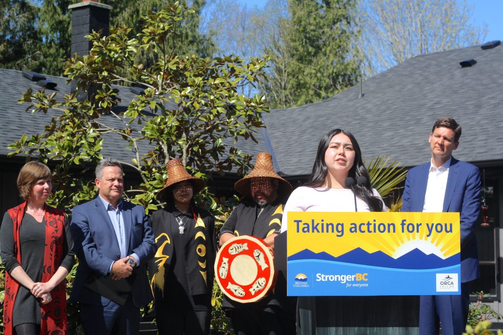 A young woman with long black hair speaks at a podium that reads Taking Action for You