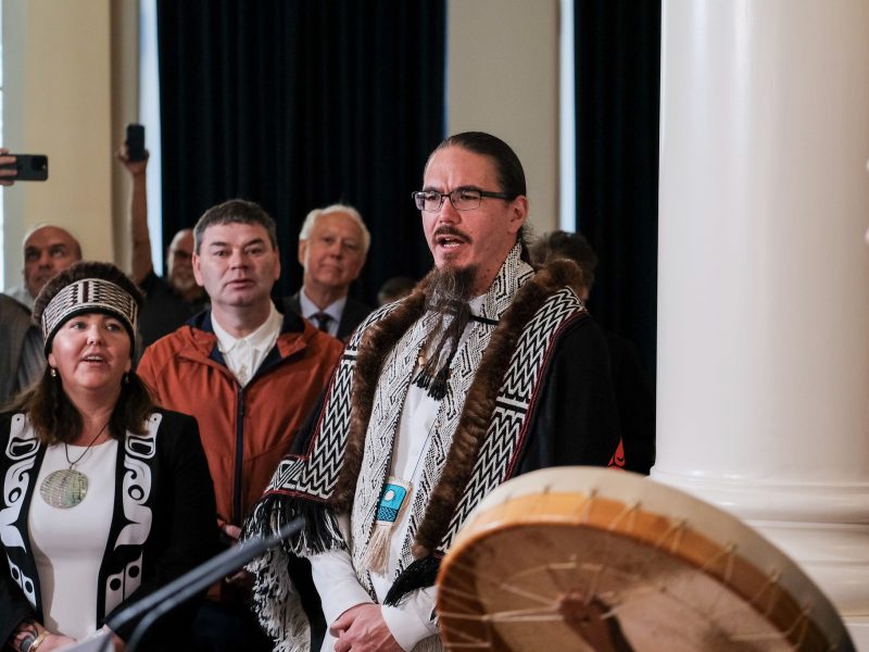 President of the Haida Nation Gaagwiis wears glasses and in Haida regalia sings in a group of people that include drummers and Minister Murray Rankin
