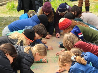 Students and adults kneel around a long brown piece of paper laid on the ground and colour on it with crayons.