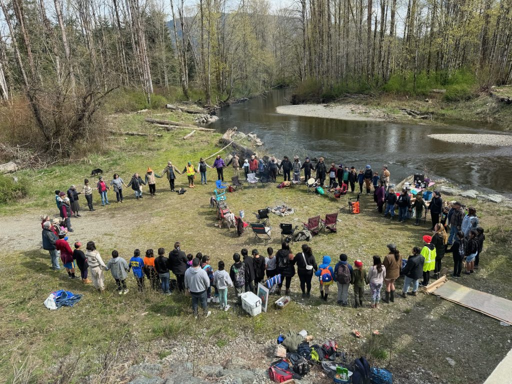 A birds-eye-view photo of community members gathered in a large circle, holding hands, on the banks of the river.