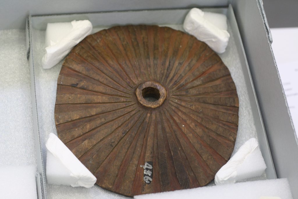 A wooden spindle whorl with radiating lines and a central hole lies in a white museum box