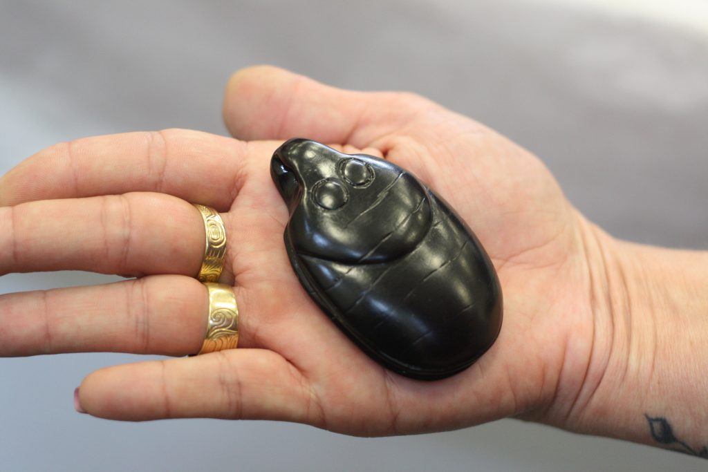 A black pendant in the shape of a beetle is cradled in a person's hand with gold rings on the hand