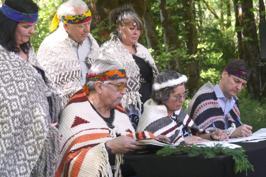 Lyackson Chief Pahalicktun (Richard Thomas), Cowichan Tribes Chief Cindy Daniels (Sulsulxumaat) and Premier Eby sign the agreement. They are sitting at a table, blanketed, while three witnesses stand behind them.