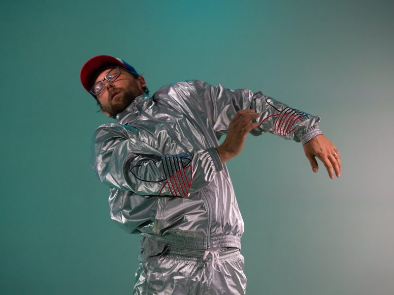 A man in a silver suit and ball cap dances in front of a green background