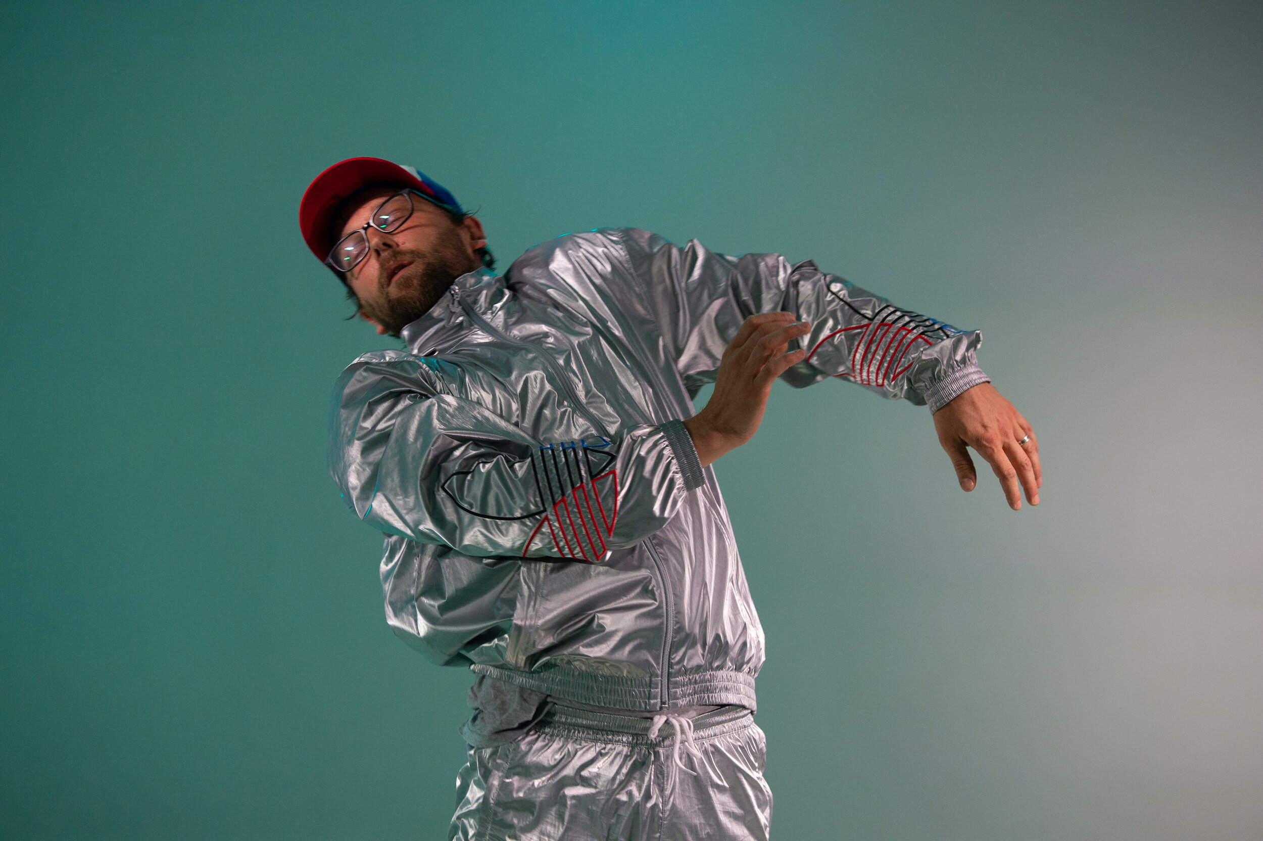 A man in a silver suit and ball cap dances in front of a green background