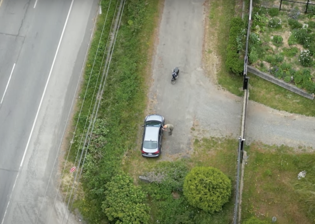 Aerial view of a sedan with a motorcycle parked about 10 metres away. A man stands leaning over the window of the car and appears to be recording or taking photos of the people in the car.