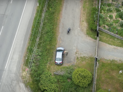 Aerial view of a sedan with a motorcycle parked about 10 metres away. A man stands leaning over the window of the car and appears to be recording or taking photos of the people in the car.
