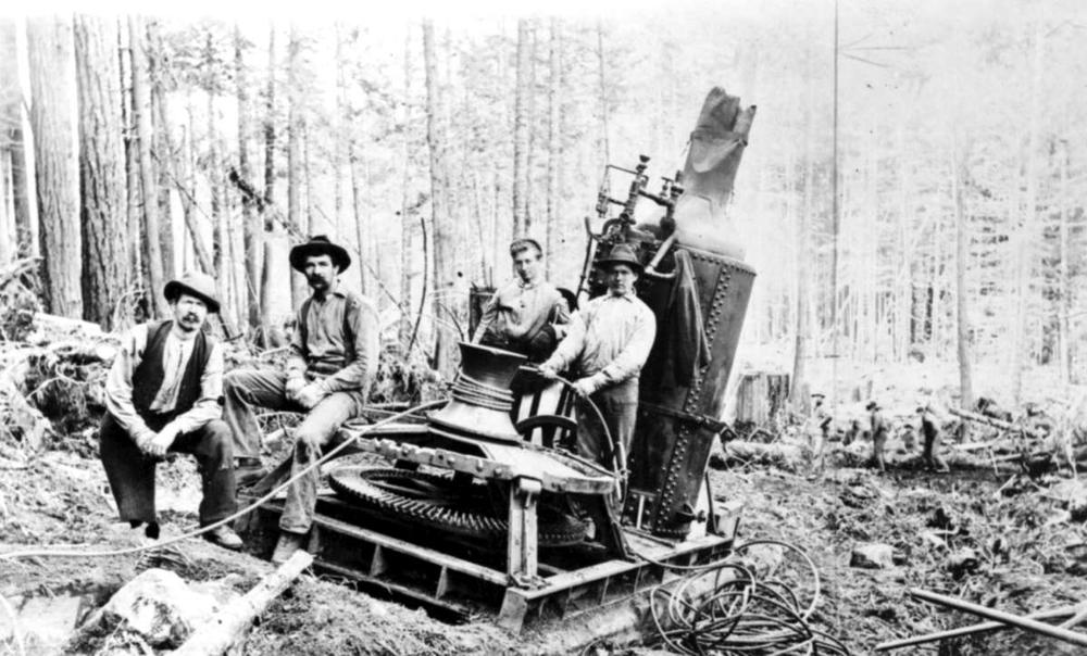 Black and white historical photo shows one of the first steam donkey machines in B.C. in a forest with three men posing on it