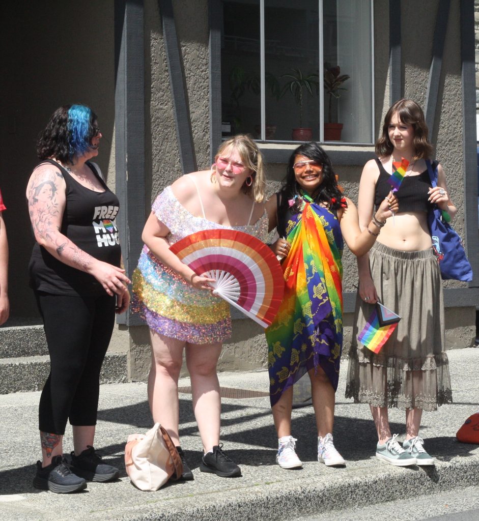 Four people stand on the side of the road dressed up in pride colours, one holds an umbrella with the lesbian pride colours of orange, pink and white.