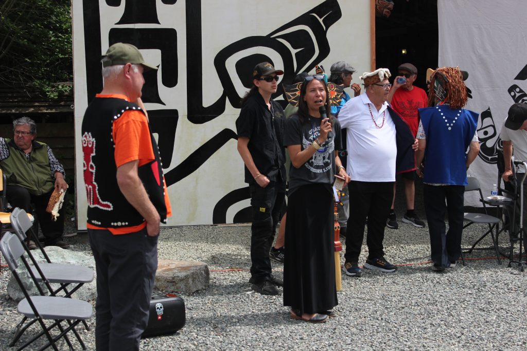 An Indigenous woman in a black skirt speaks into a microphone. Next to her are a group of Elders in regalia and one younger person. In the background are decorative blankets.