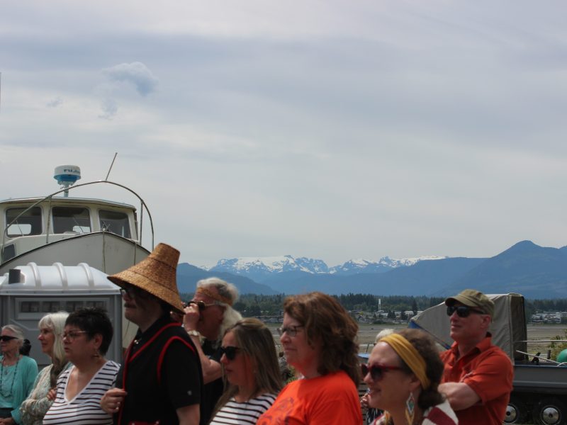 A group of people stand in the foreground looking in the same direction. One tall person wears a traditional cedar-woven hat. The K'ómoks Glacier is in the background.