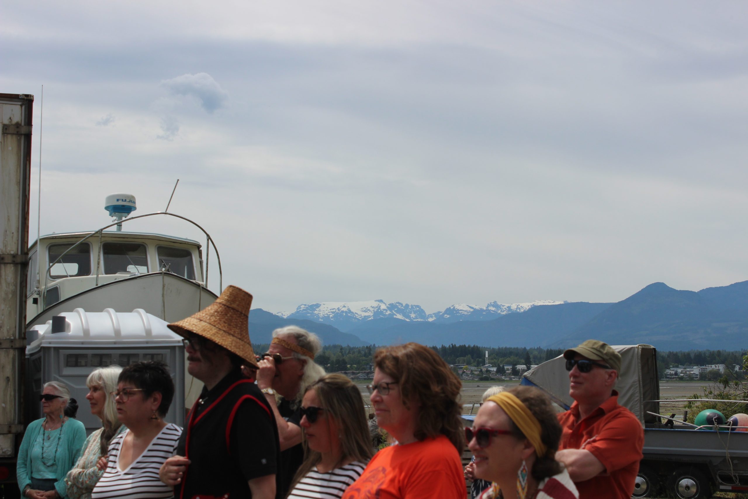 A group of people stand in the foreground looking in the same direction. One tall person wears a traditional cedar-woven hat. The K'ómoks Glacier is in the background.