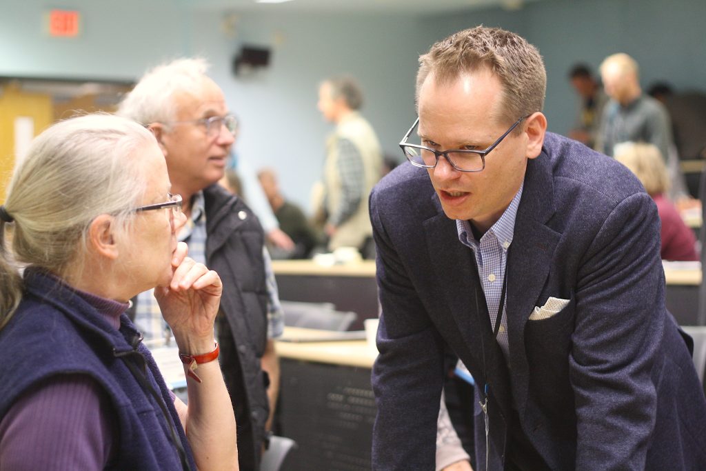 A man in a suit and glasses leans down to speak to a grey haired woman who is listening intently to 6PPD-q