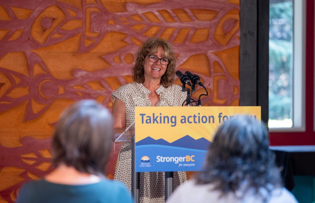 A woman with curly blond hair and black glasses smiles at a podium.