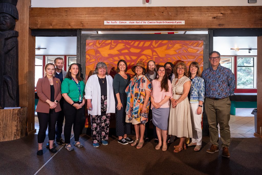 A group of men and women stand smiling in front of a backdrop of Indigenous art.